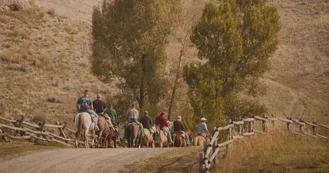 A group of people ride horses along a dirt path near a wooden fence.