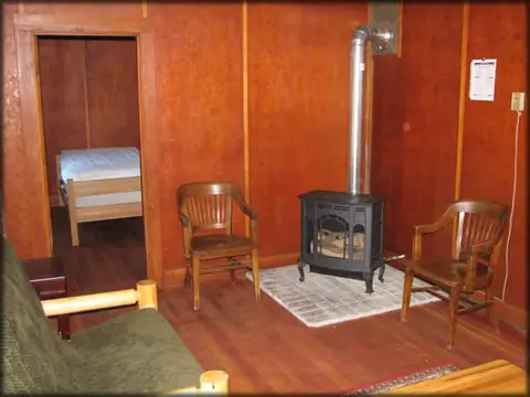 An interior view of a room with wooden walls, a wood stove, and two chairs.