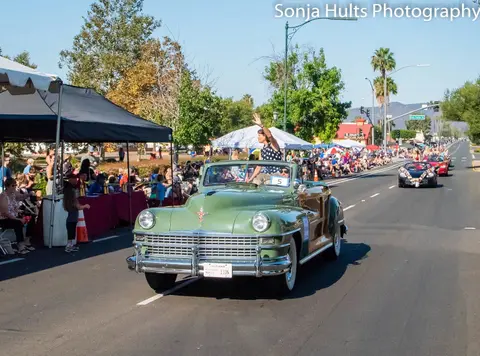 A vintage green convertible drives down a street in a parade, with people waving from the vehicle and crowds watching along the side.