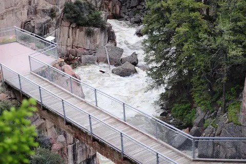 A wooden footbridge spans a rushing river through a rocky canyon.