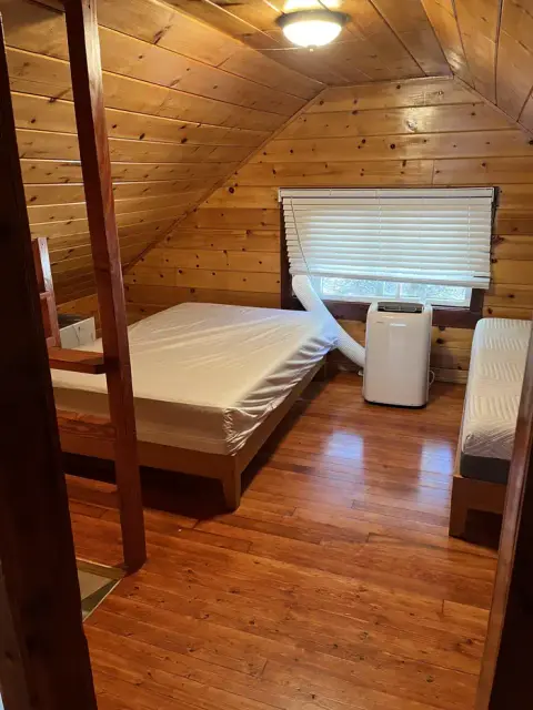 An attic bedroom with wooden walls and ceiling containing beds and a portable air conditioner.