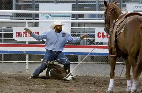 A cowboy wrestles a calf in an arena while a horse stands nearby.