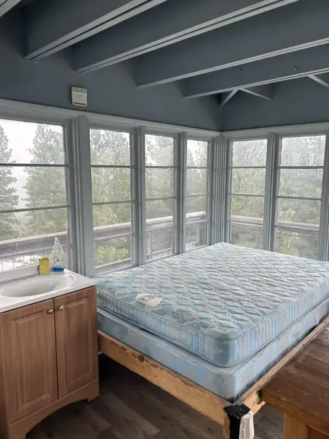 An interior view of a room with a bed and a sink, surrounded by large windows overlooking trees.