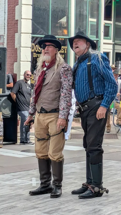 Two men dressed in historic western attire stand on a stage outdoors.