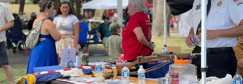 People gather around a booth displaying flags and a rifle outdoors.