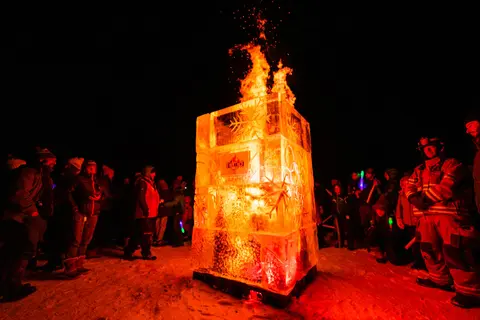 A large ice sculpture with fire burning inside and on top, surrounded by a crowd at night.