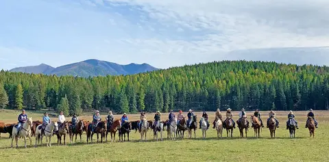 A group of horseback riders in a grassy field with a forest and mountains in the background.