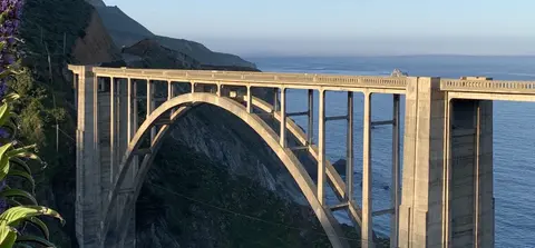 A large concrete arch bridge spans a coastal canyon above the ocean.