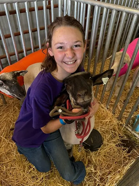 A smiling young person holds a sheep inside a pen with straw bedding.