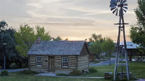 An old log cabin with a wooden windmill standing beside it at sunset.