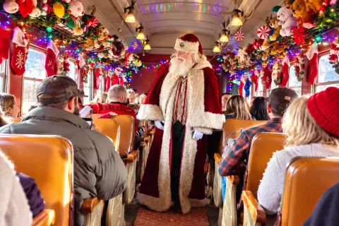 Santa Claus walks down the aisle of a festively decorated train car filled with passengers.