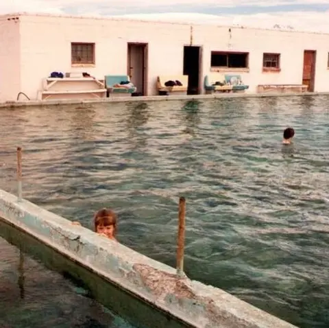 People swim in an outdoor pool in front of a long white building.