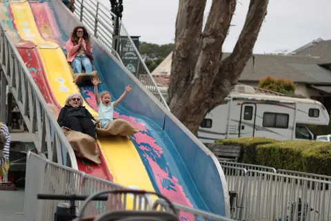 People ride down a large multi-lane slide sitting on burlap sacks.