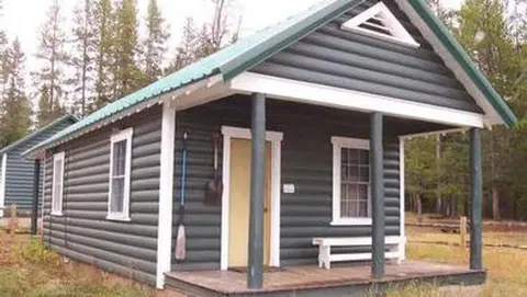 A small dark log cabin with a green metal roof and front porch stands in a wooded area.