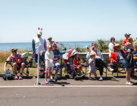 A crowd of people watch a parade near the ocean on a sunny day.