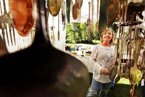 A woman stands inside a craft booth surrounded by hanging silverware displays.