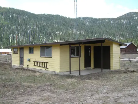 A single-story yellow building with a covered porch and carport sits in a landscape with pine-covered mountains.