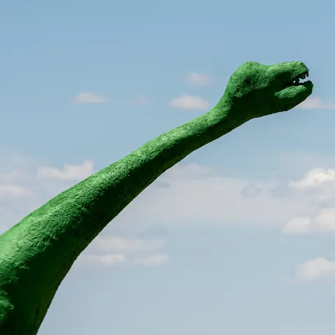 A green dinosaur statue neck and head against a blue sky with clouds.