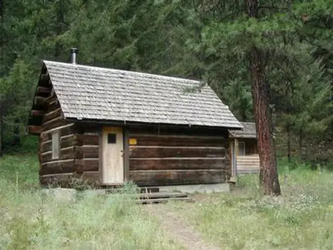 A rustic log cabin sits surrounded by trees and tall grass.