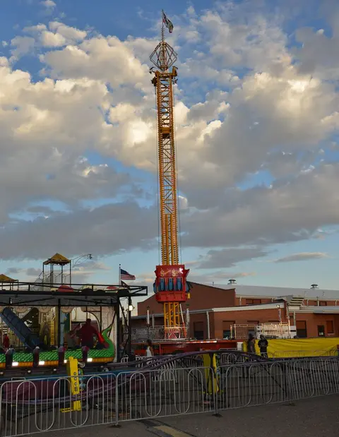 A tall drop tower ride stands at an outdoor fair under a cloudy sky.