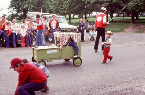 People gather outdoors around a small wooden cart and a child in cowboy attire.