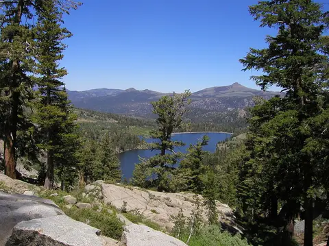 A scenic view of a mountain lake framed by pine trees and rocky terrain under a clear blue sky.