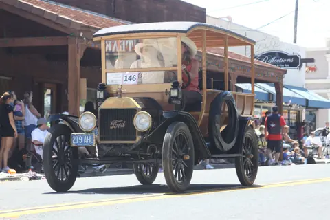 A vintage Ford automobile drives down a street during an event.