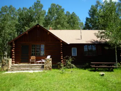 A log cabin stands in a clearing surrounded by trees under a blue sky.