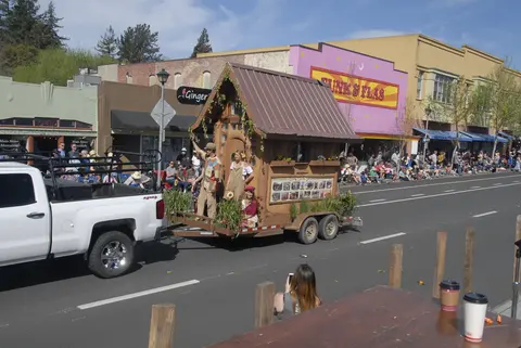 A parade float designed like a wooden cottage is towed by a pickup truck down a street lined with spectators.