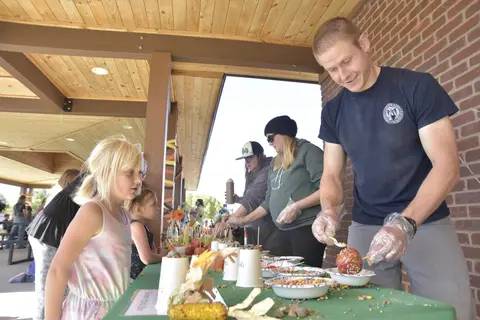 A man wearing gloves prepares caramel apples at a table where children wait.