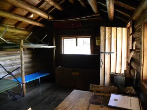 An interior view of a rustic log cabin with cots and a wooden table.