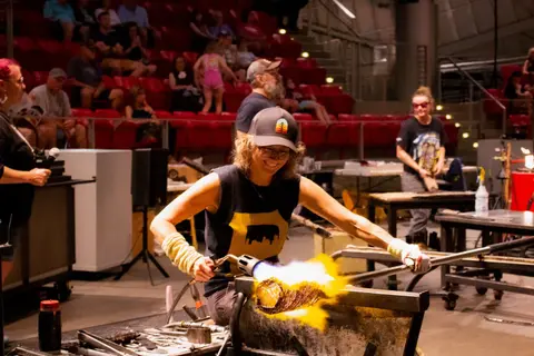 A woman works with hot glass using a torch in front of an audience.