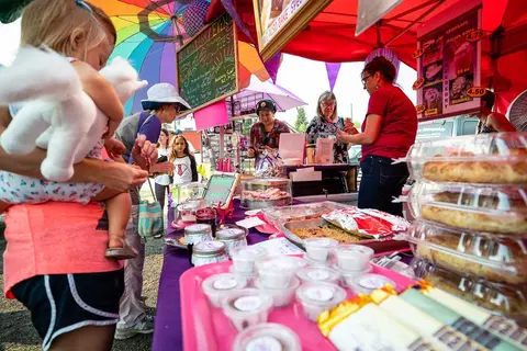 People at a food booth selling pies and other baked goods under colored tents.