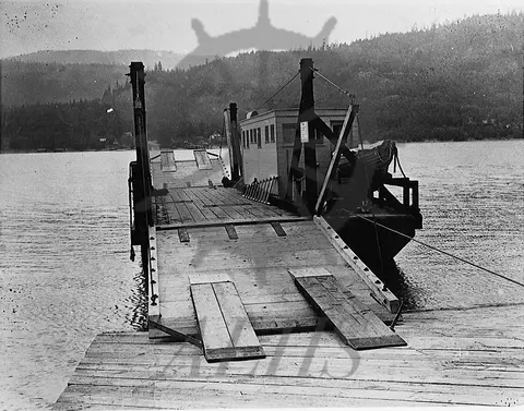 A historic black and white photograph of a ferry docked at a wooden pier.