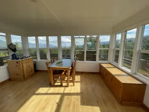 An interior shot of a wooden cabin room surrounded by windows offering a mountain view.