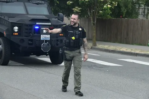A smiling officer in uniform directs traffic in front of an armored vehicle.