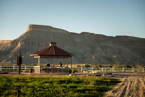 A gazebo stands in front of a large mountain and white fence under a clear sky.