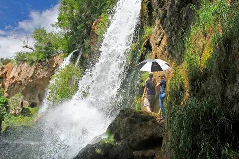 Two people stand near a waterfall holding an umbrella.