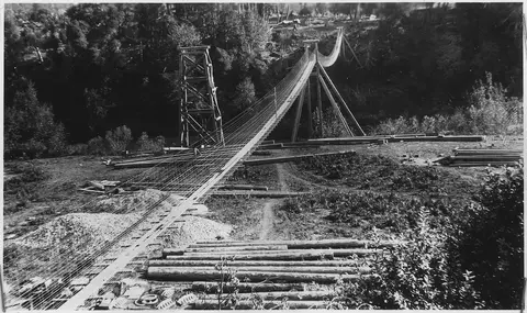 An old black and white photo shows a suspension bridge under construction.