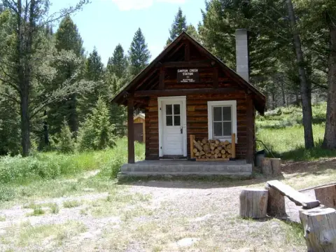 A rustic log cabin sits in a forested area with a sign above the door.