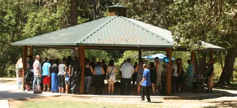 A group of people gathers under a green-roofed pavilion in a park.