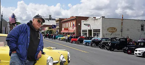 A man leans against a classic car on a street lined with vintage automobiles and historic buildings.