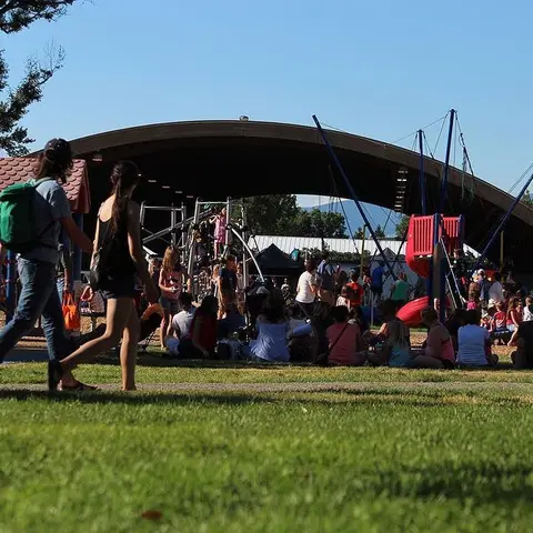 People walking and gathering at a public park near a playground and large pavilion.