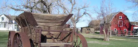 An old wooden cart sits on a grassy field near a red barn and other historic buildings.