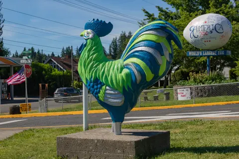 A colorful rooster statue stands on a concrete pedestal near a street.