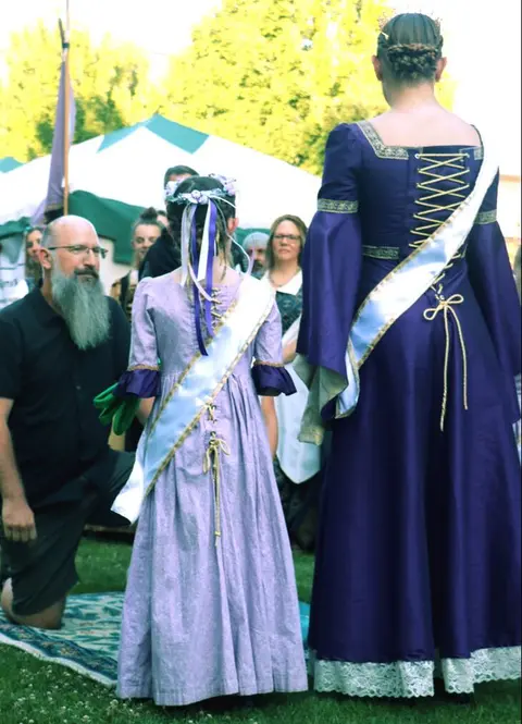 A man kneels before people in period costumes outdoors.