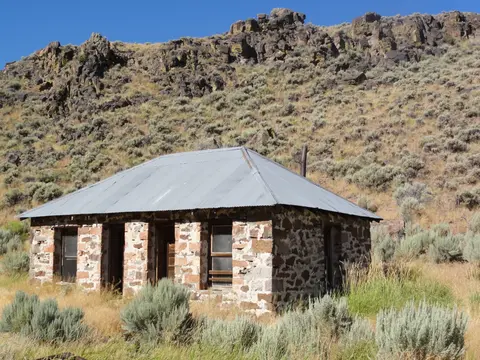 A stone building with a metal roof sits at the base of a rocky hillside.