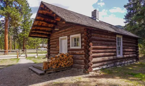 A rustic log cabin stands in a wooded area with a stack of firewood next to its porch.