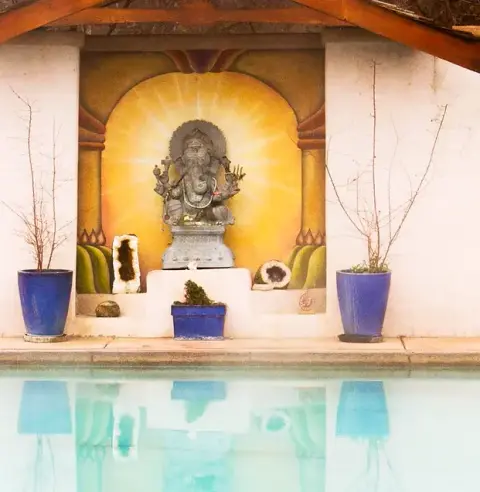 A statue of Ganesha sits on an altar with a painted mural behind it next to a swimming pool.