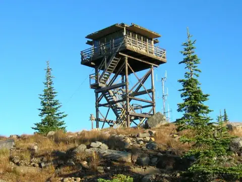 A wooden fire lookout tower stands on a rocky hill with trees under a blue sky.